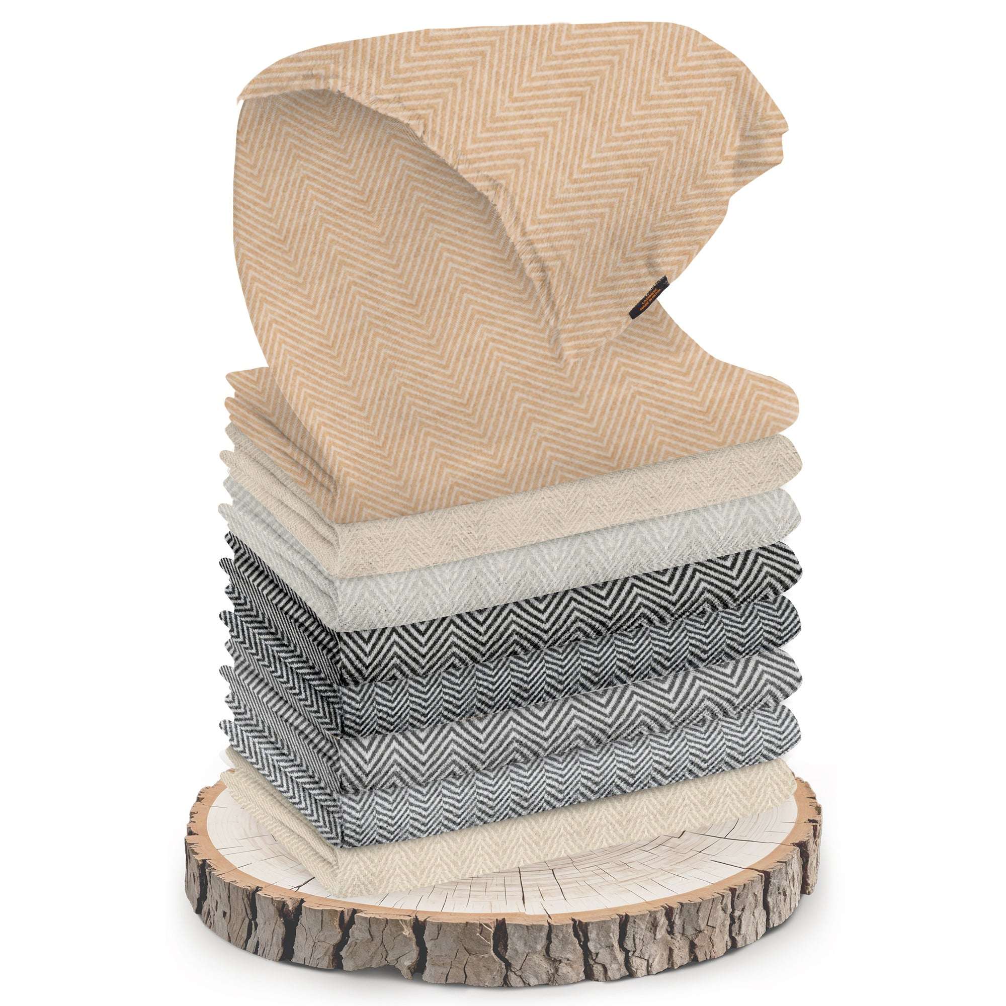 Mehrere gefaltete Schals mit Fischgrätenmuster in verschiedenen Beige-, Grau- und Schwarztönen sind auf einer Baumscheibe gestapelt. Oben ist ein goldbeiger Kaschmirschal halb ausgebreitet und gut sichtbar platziert. Die Oberfläche der Schals zeigt ein gut erkennbares, mittelgroßes Zickzackmuster. Die Baumscheibe hat eine sichtbare Rinde und Baumschutzringe. Der Hintergrund ist weiß und neutral gehalten, nichts Weiteres ist sichtbar.
