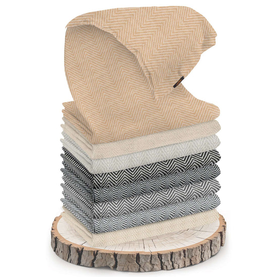 Mehrere gefaltete Schals mit Fischgrätenmuster in verschiedenen Beige-, Grau- und Schwarztönen sind auf einer Baumscheibe gestapelt. Oben ist ein goldbeiger Kaschmirschal halb ausgebreitet und gut sichtbar platziert. Die Oberfläche der Schals zeigt ein gut erkennbares, mittelgroßes Zickzackmuster. Die Baumscheibe hat eine sichtbare Rinde und Baumschutzringe. Der Hintergrund ist weiß und neutral gehalten, nichts Weiteres ist sichtbar.