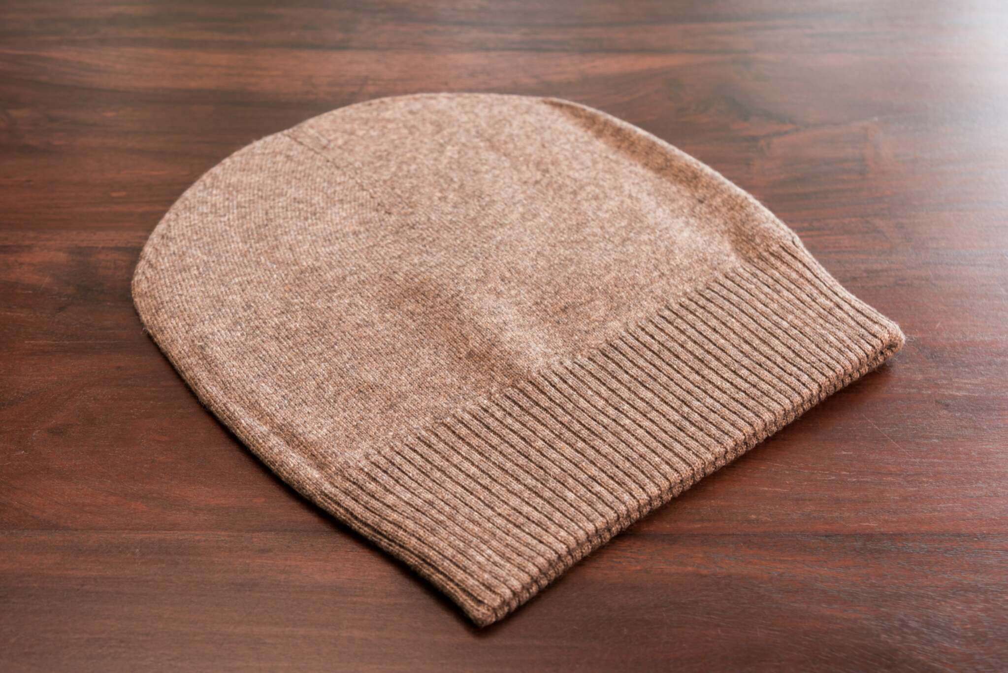 Auf dem Bild liegt eine glatte, dunkelbraune Kaschmir-Beanie mit breitem, geripptem Umschlag auf einer Holzoberfläche.