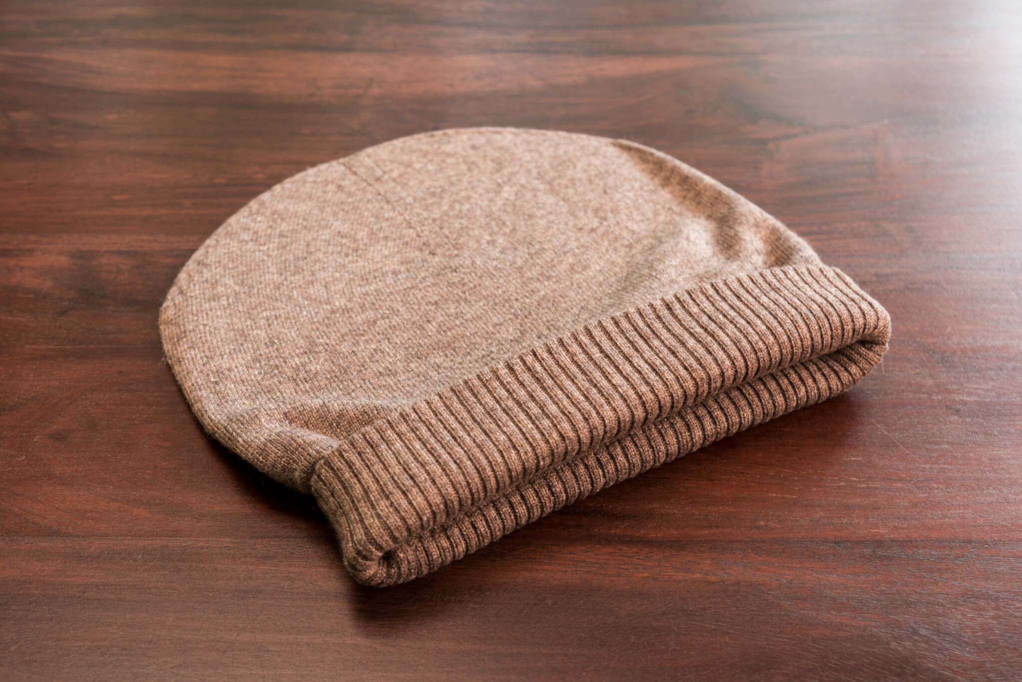 Auf dem Bild ist eine einzelne dunkelbraune Kaschmirmütze im Beanie-Stil zu sehen. Die Mütze ist glatt gestrickt mit einem breiten, gerippten Umschlag am unteren Rand. Sie liegt flach auf einer glatten, braunen Holzoberfläche. Das Licht betont die feine Textur des Kaschmirstoffs. Sonstige Gegenstände oder Muster sind nicht sichtbar.
